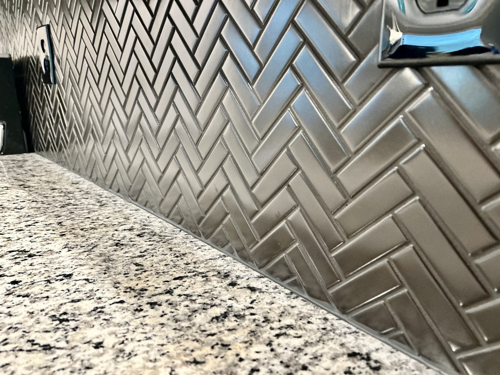 Metallic gray herringbone tile backsplash installed above a granite countertop